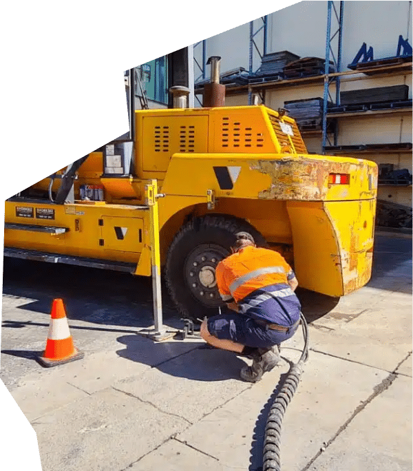 A technician in high-visibility workwear servicing a large industrial yellow machine tire with specialized tools at a workshop.