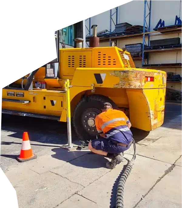 A technician in high-visibility workwear servicing a large industrial yellow machine tire with specialized tools at a workshop.